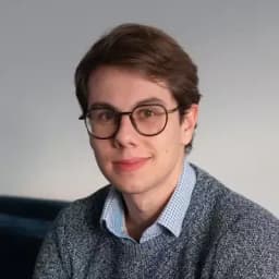 Jeune homme aux cheveux bruns courts, portant des lunettes rondes et une chemise bleue, souriant légèrement devant un fond neutre